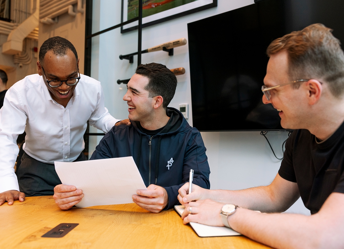 Three colleagues smiling and discussing documents together at a meeting table.