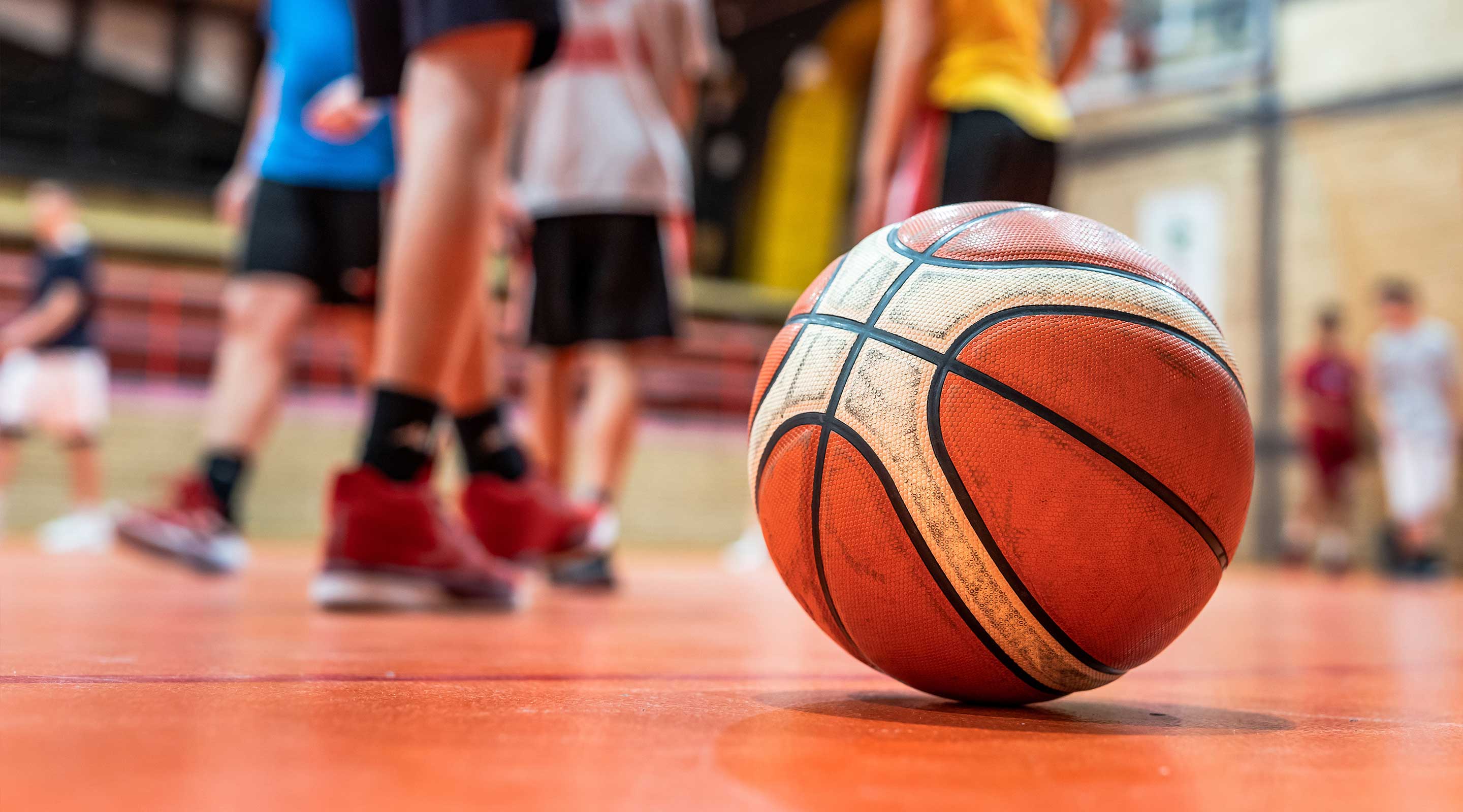 Basketball on a gym floor with players standing in the background.